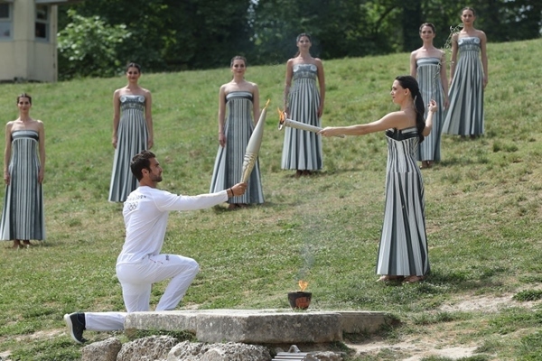 扮演最高女祭司的演員瑪麗·米娜（前右）點(diǎn)燃第一棒火炬手——希臘運(yùn)動(dòng)員、東京奧運(yùn)會(huì)男子賽艇項(xiàng)目金牌得主斯特凡諾斯·恩圖斯科斯手中的火炬。新華社記者 趙丁喆 攝