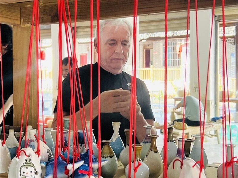 鄉(xiāng)愁如同一根根紅線，將游子與故鄉(xiāng)永遠(yuǎn)地牽連到一起?；逄账囄幕瘎?chuàng)意一條街的母氏陶藝館內(nèi)，紅線吊著陶瓶，思緒游走萬(wàn)里。摩爾多瓦“信息標(biāo)簽”通訊社社長(zhǎng)亞歷山大·塔納斯端詳著館內(nèi)展示的陶藝作品，并挑選出心儀的小擺件帶回摩爾多瓦，也帶走花茂村的“鄉(xiāng)愁”。（張亞欣攝）